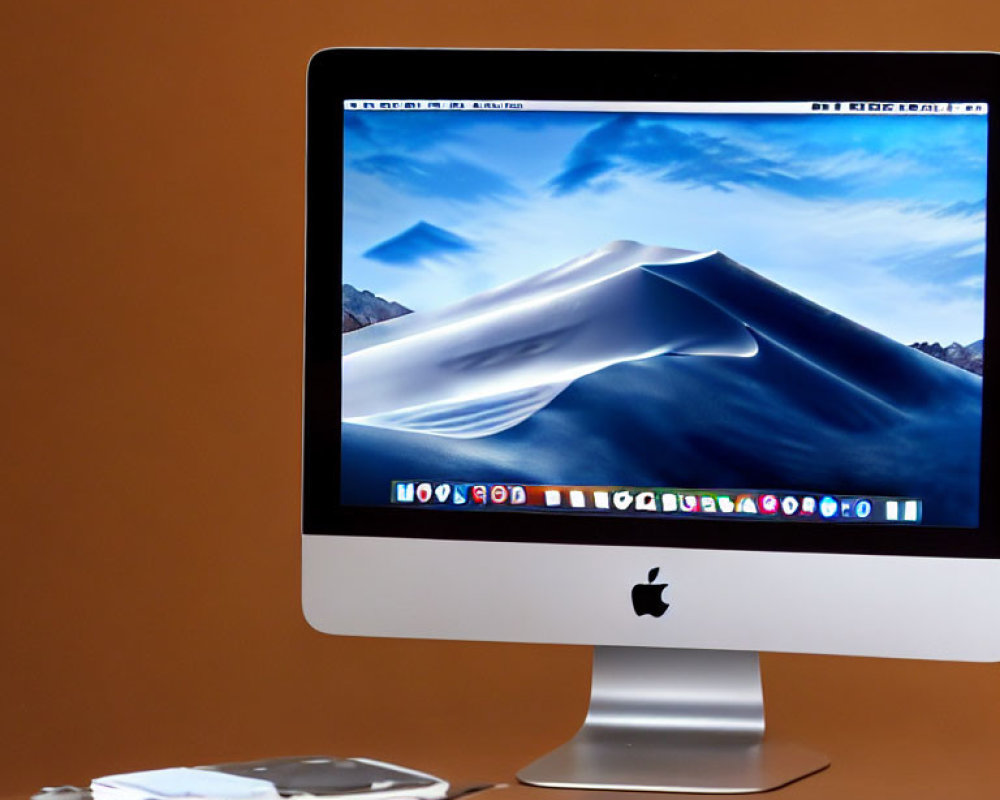 Desktop setup with iMac, desert dune wallpaper, smartphone, and glasses on brown desk