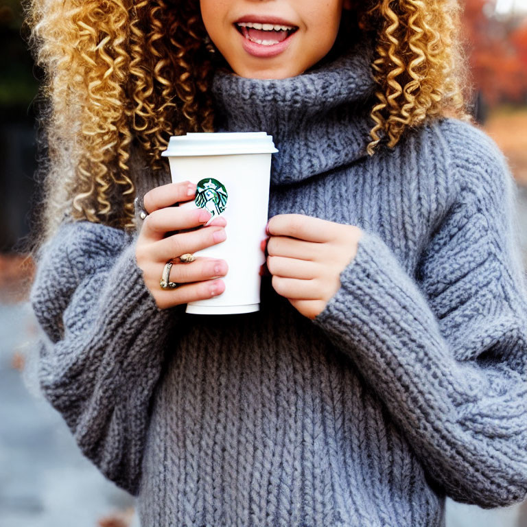 Curly-haired person in grey sweater with Starbucks cup and rings.