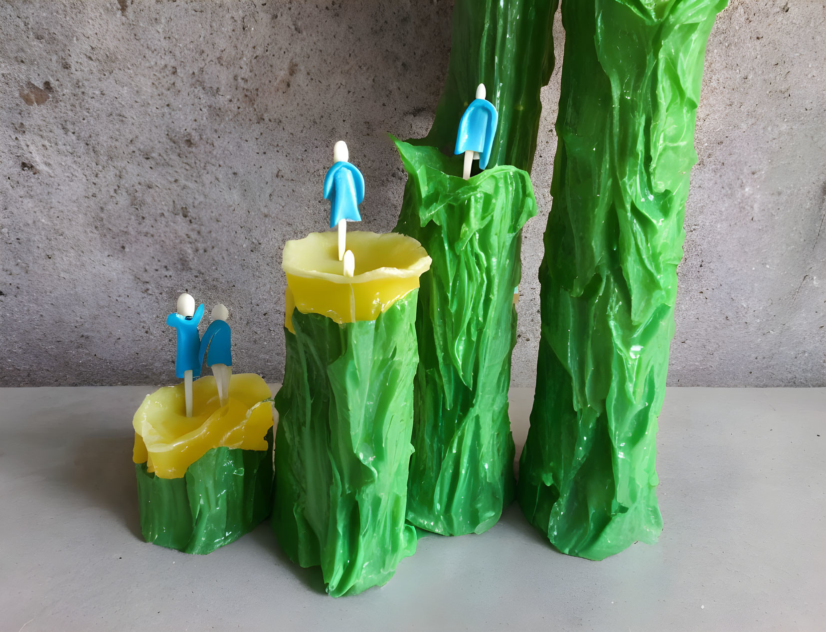 Three green textured candles with blue figurines on concrete background