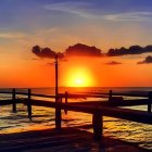 Ocean sunset viewed from pier with railing silhouette and sparse trees against orange sky.