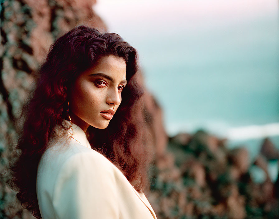 Curly-haired woman in beige outfit glances sideways against rocky terrain and blurred seascape.