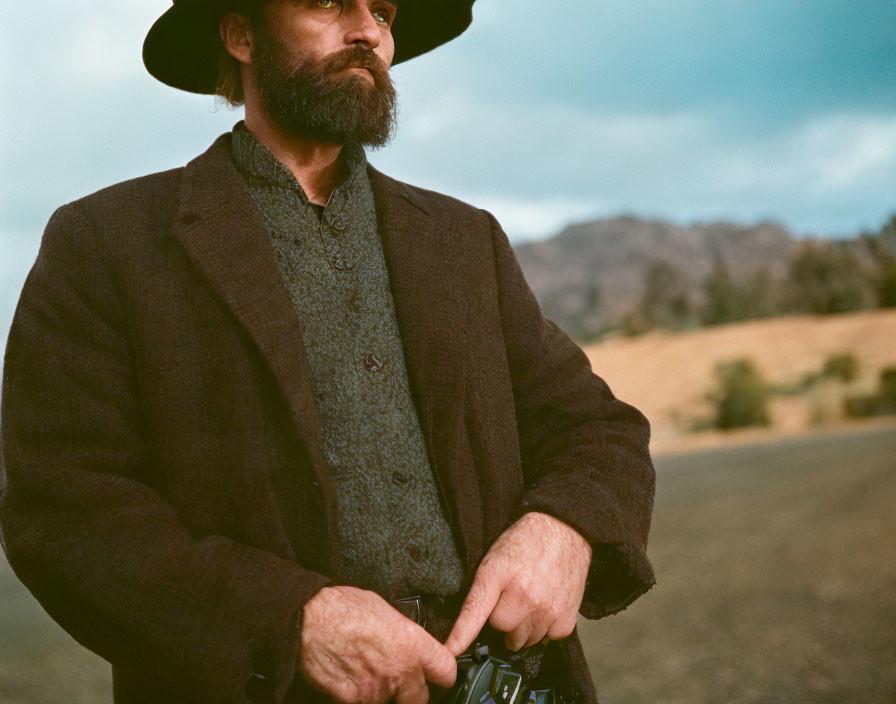 Bearded man in hat and jacket against scenic backdrop.