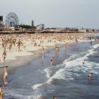 Crowded beach with swimmers and sunbathers, boardwalk with amusement park rides, clear blue