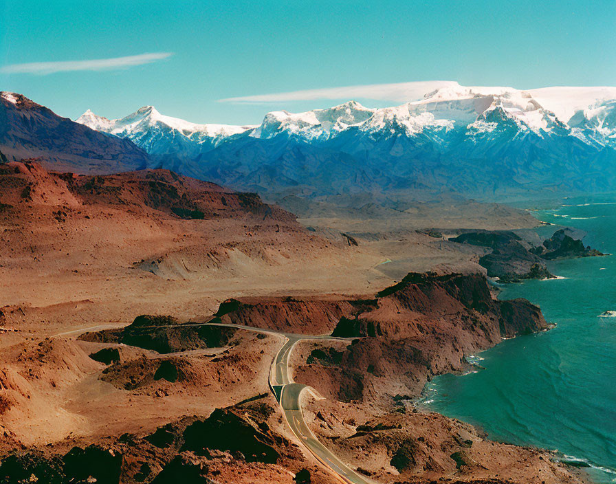 Scenic winding road through barren landscape, coastline, snow-capped mountains.