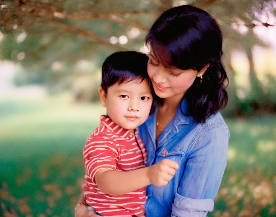 Woman embracing young boy under tree in denim and red-striped tee