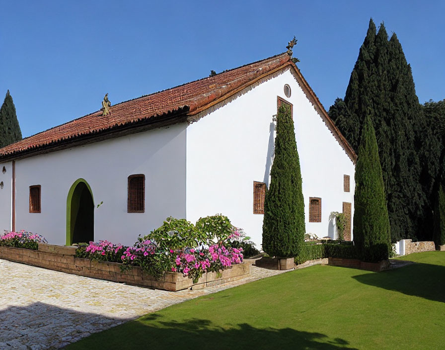 One-story white house with clay roof tiles and green arched door amidst pink flowers and tall conifer