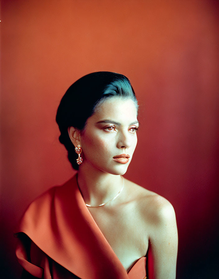 Dark-haired woman in red off-the-shoulder dress with red lipstick and large earrings against red backdrop