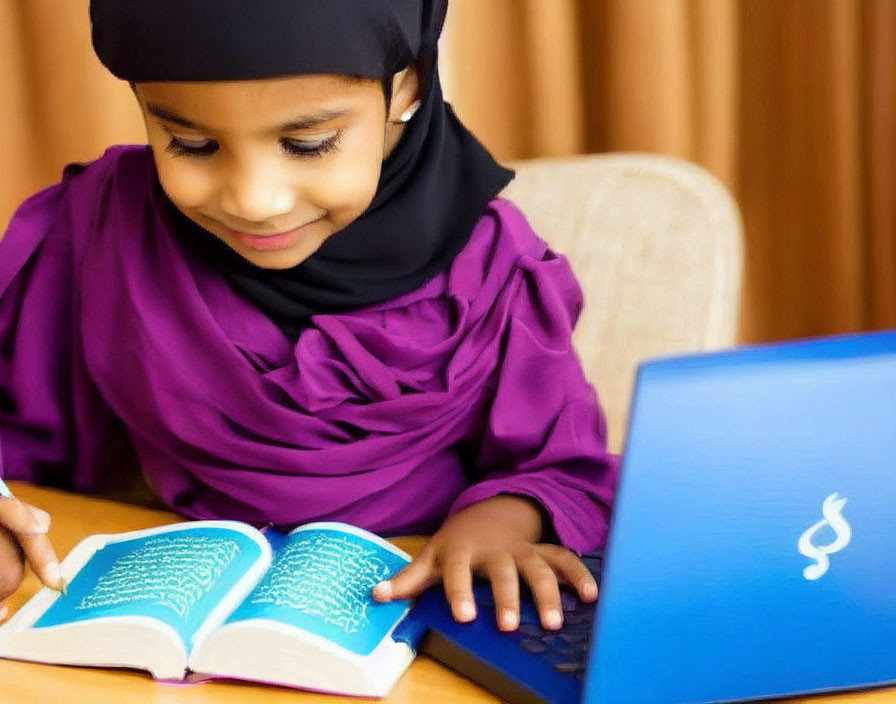 Young girl in hijab reading book and using laptop on desk