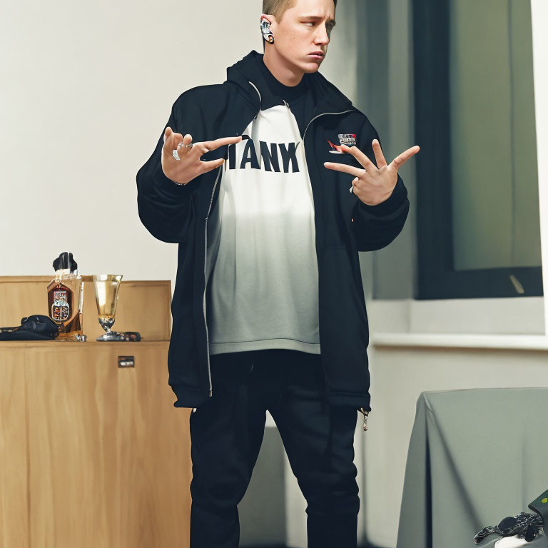 Person in Black Jacket and White T-Shirt with "TANK" Text Making Hand Gesture in Room with