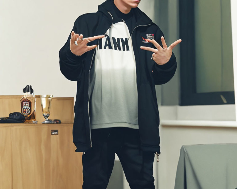 Person in Black Jacket and White T-Shirt with "TANK" Text Making Hand Gesture in Room with