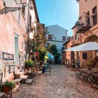 Vibrant painting of medieval village with characters, buildings, river, boats, and plane