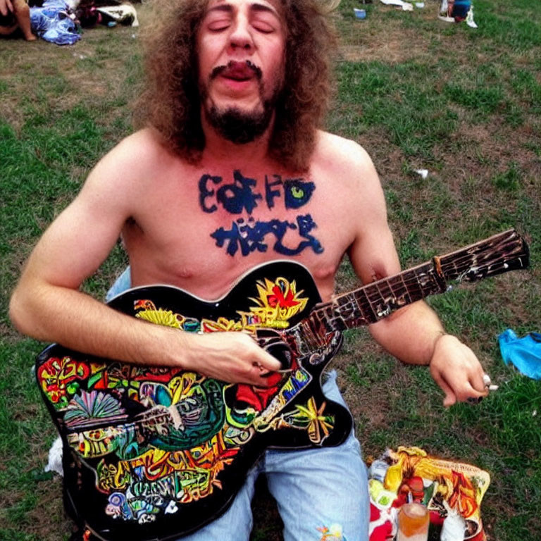 Curly-Haired Man Playing Colorful Guitar Outdoors in Grass with Painted Words on Chest