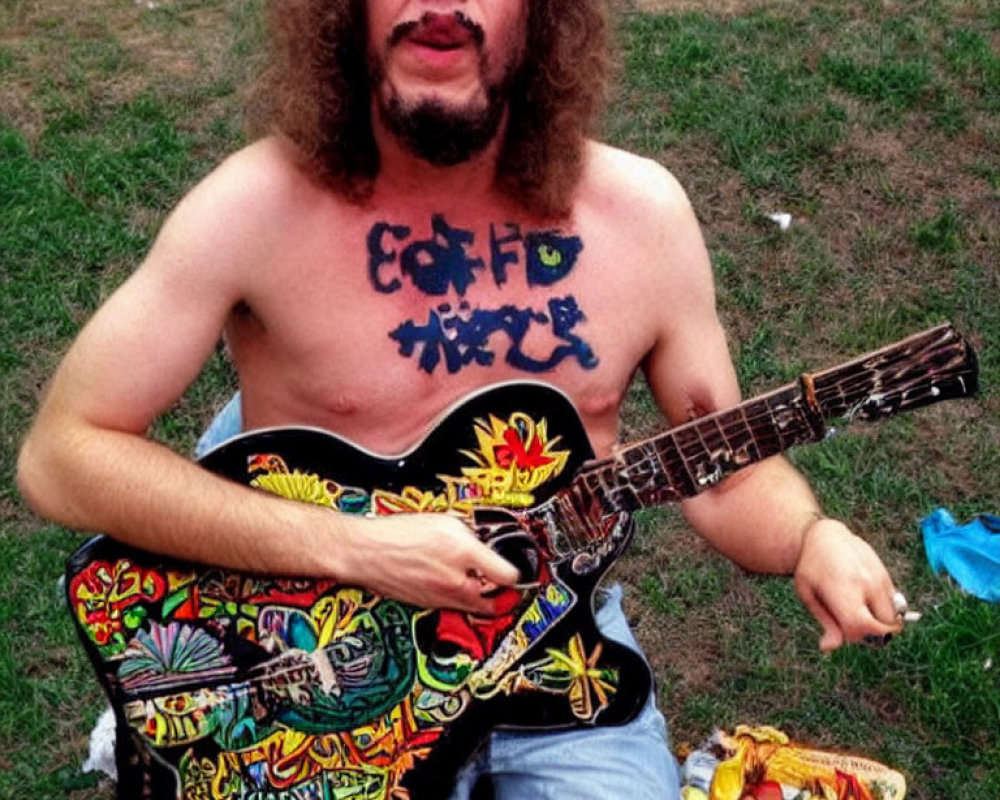 Curly-Haired Man Playing Colorful Guitar Outdoors in Grass with Painted Words on Chest