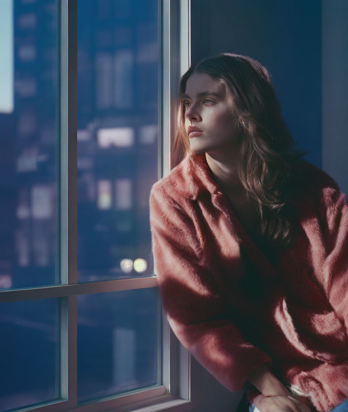 Long-haired person in pink sweater gazes out window at city twilight.