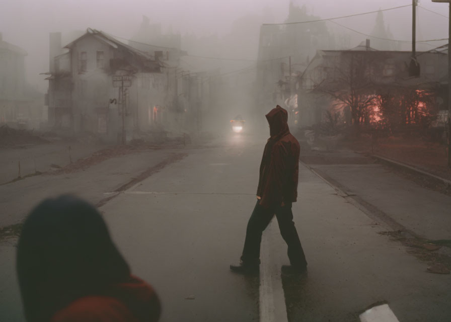 Person in red hoodie walking on misty street with run-down houses and approaching car headlights.