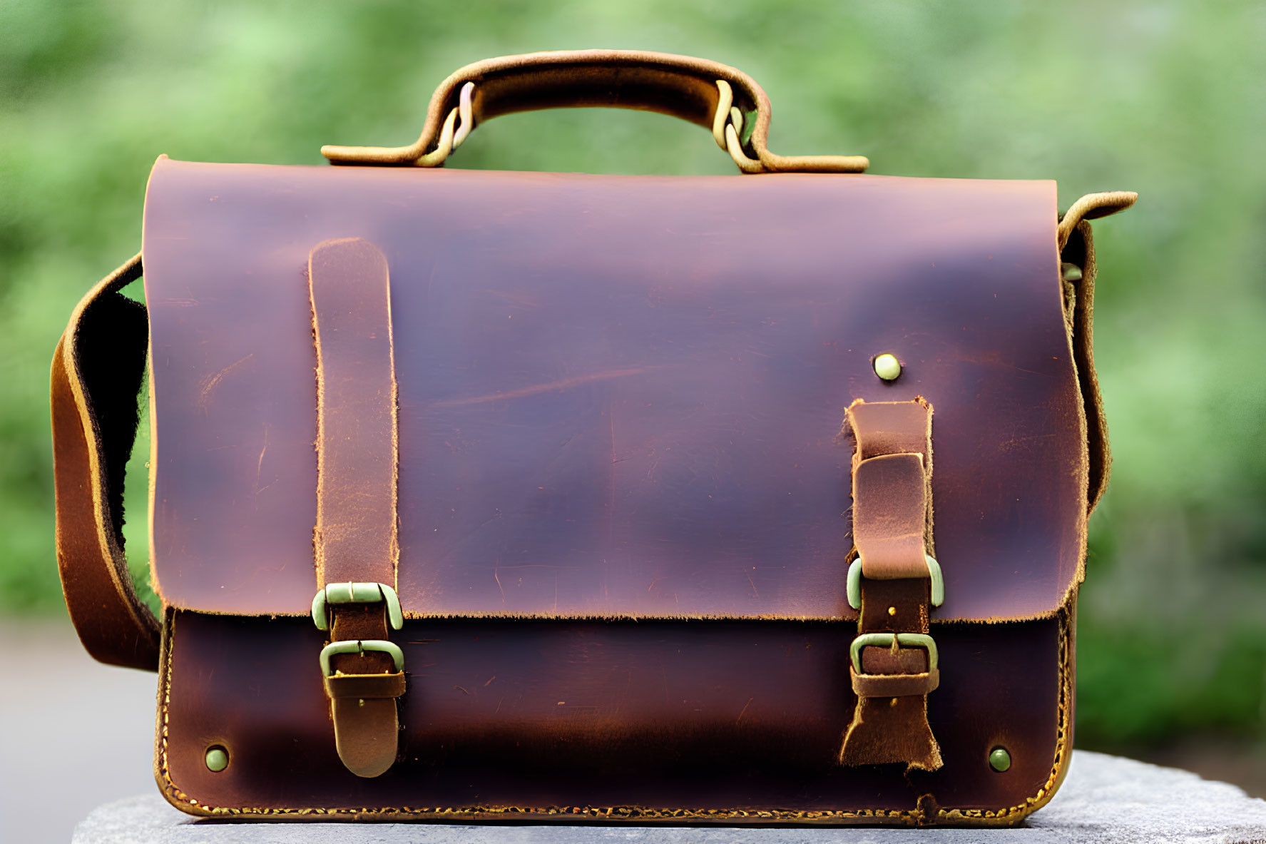 Brown Leather Satchel with Front Strap Closure and Brass Buckles on Blurred Green Background