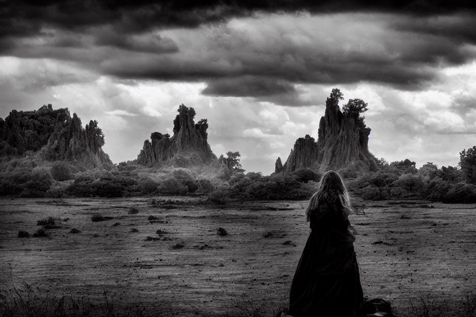 Monochrome image of cloaked figure in barren landscape with towering rocks under dramatic sky