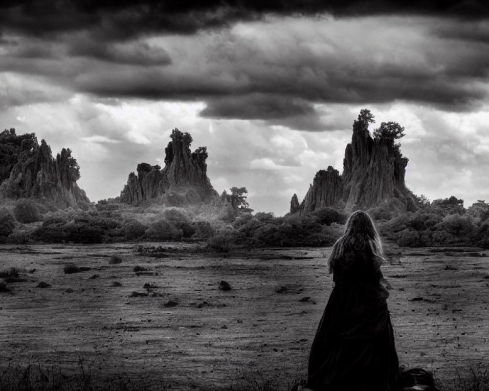 Monochrome image of cloaked figure in barren landscape with towering rocks under dramatic sky