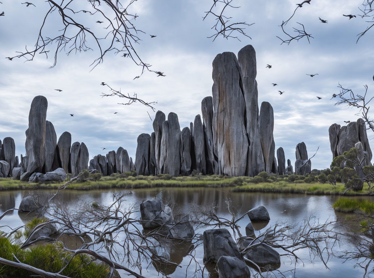 Weathered rock formations towering over serene water with scattered rocks and birds in cloudy sky