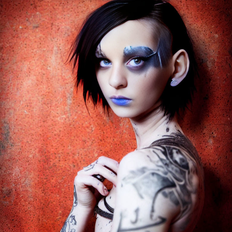 Portrait of person with dark hair, silver eye makeup, blue lipstick, and tattoos on orange background