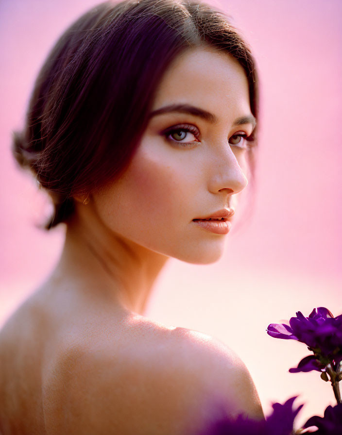 Woman with shoulder-length hair and subtle makeup on pink background with blurred purple flower