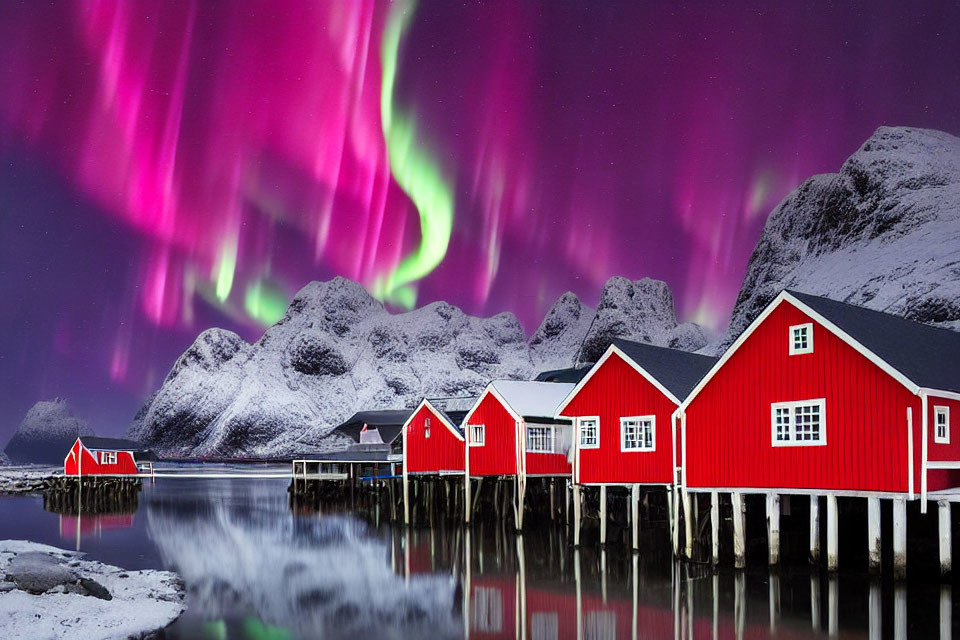 Northern Lights illuminate red cabins over lake and snowy mountains.