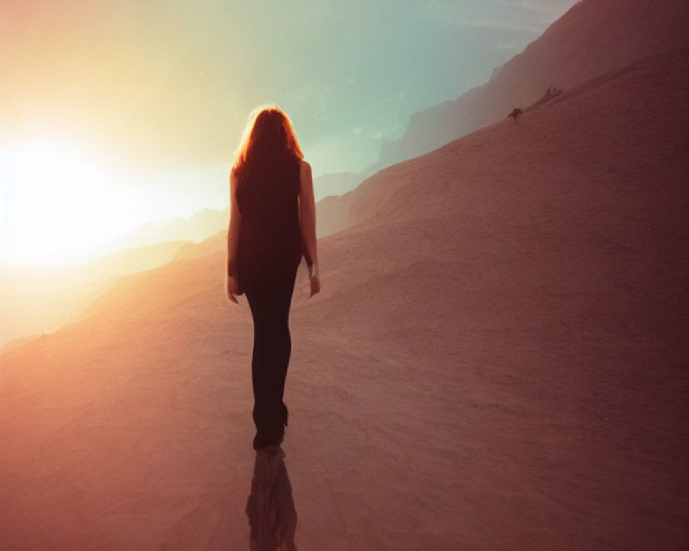 Person with long hair walking up desert sand dune at sunset