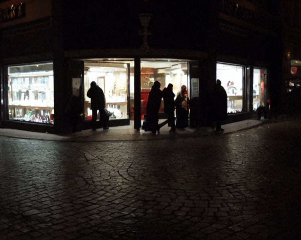 Night scene: Silhouettes passing by brightly lit store on cobblestone pavement