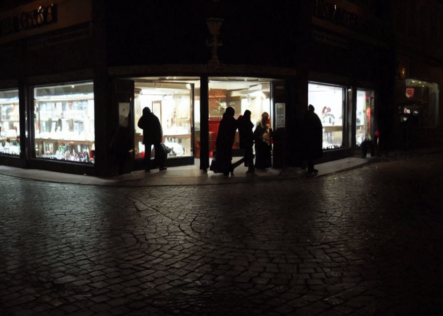 Night scene: Silhouettes passing by brightly lit store on cobblestone pavement