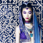 Elaborately dressed woman with blue headdress and purple outfit against patterned backdrop