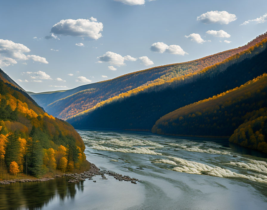 Autumnal trees along serene river in mountainous valley under bright sky.