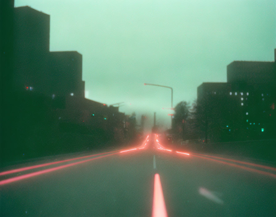 Blurred road photo with red tail light trails and silhouetted buildings at dusk or dawn