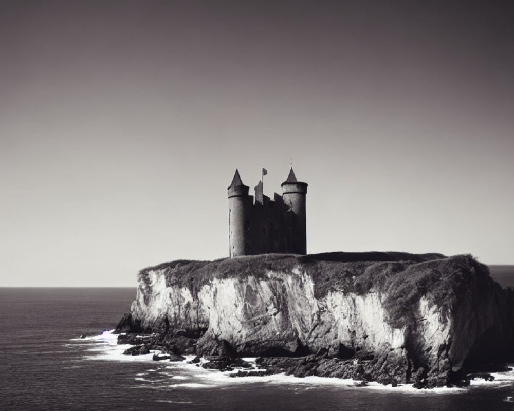 Grayscale image of castle on cliff overlooking calm sea