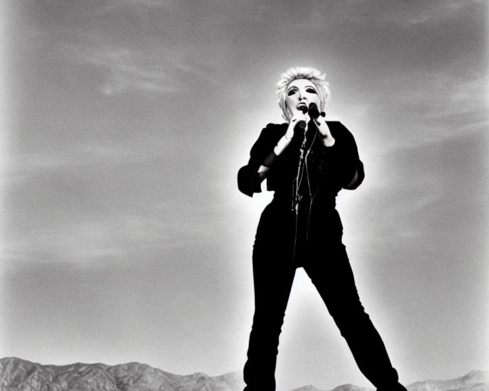 Person posing with microphone in barren landscape under dramatic sky