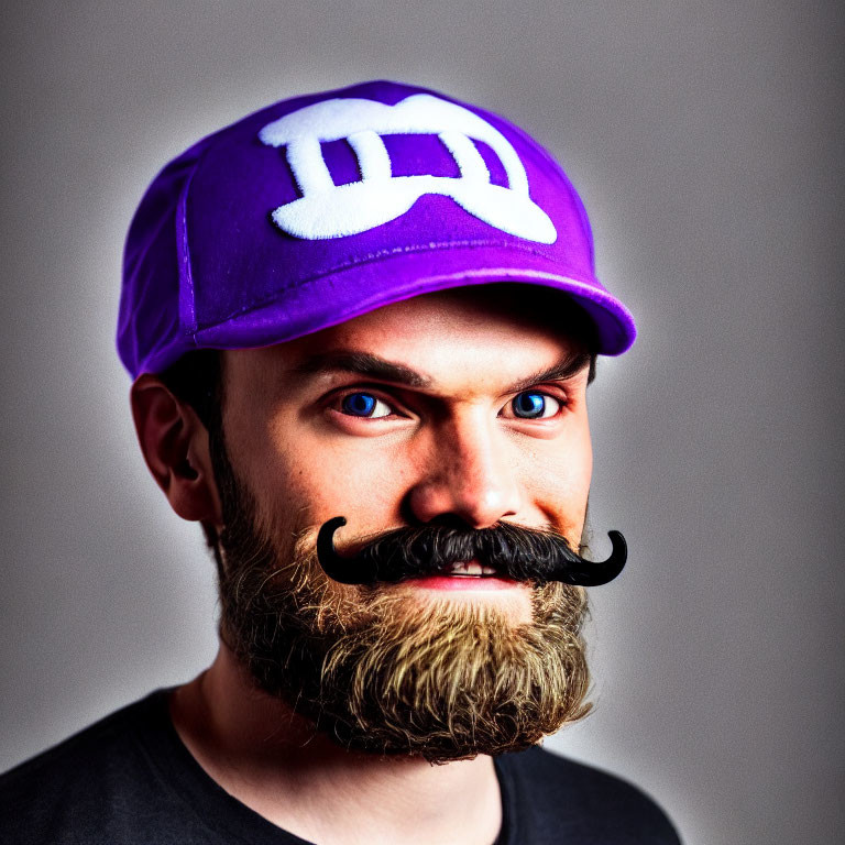 Man with Thick Beard and Whimsical Fake Mustache in Purple Cap Smiling on Grey Background