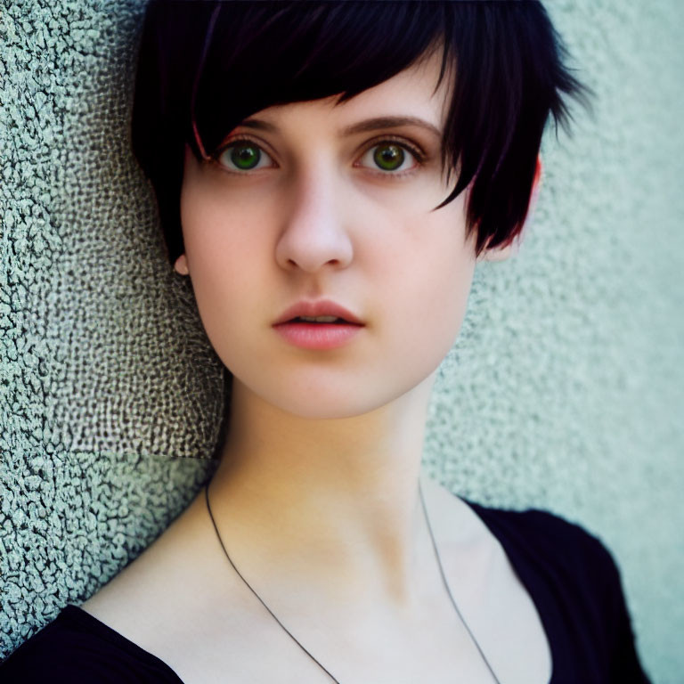 Black-haired person with green eyes in black top against textured wall.