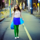 Young woman in white blouse and blue skirt stands in wet street at night with city lights in background