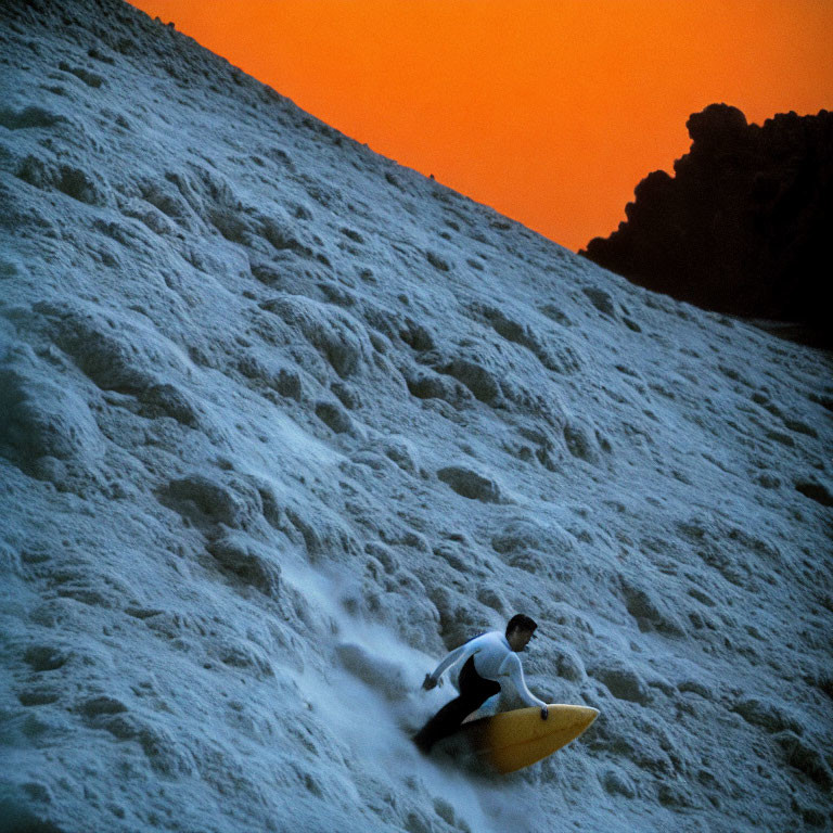 Surfer riding large wave against orange sky with foamy waters and rocks in background