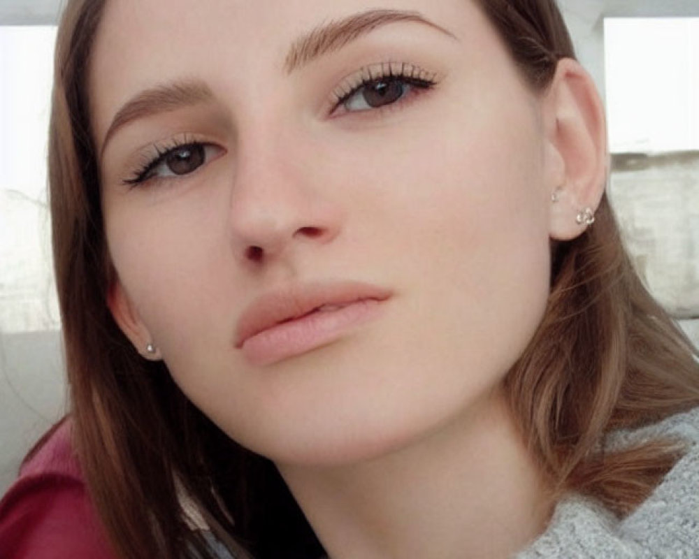 Fair-skinned woman in grey sweater takes selfie with light brown hair and makeup.