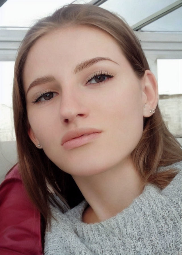 Fair-skinned woman in grey sweater takes selfie with light brown hair and makeup.