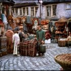 Medieval-themed market street at dusk with cloaked figures browsing stalls adorned with lights and textiles.