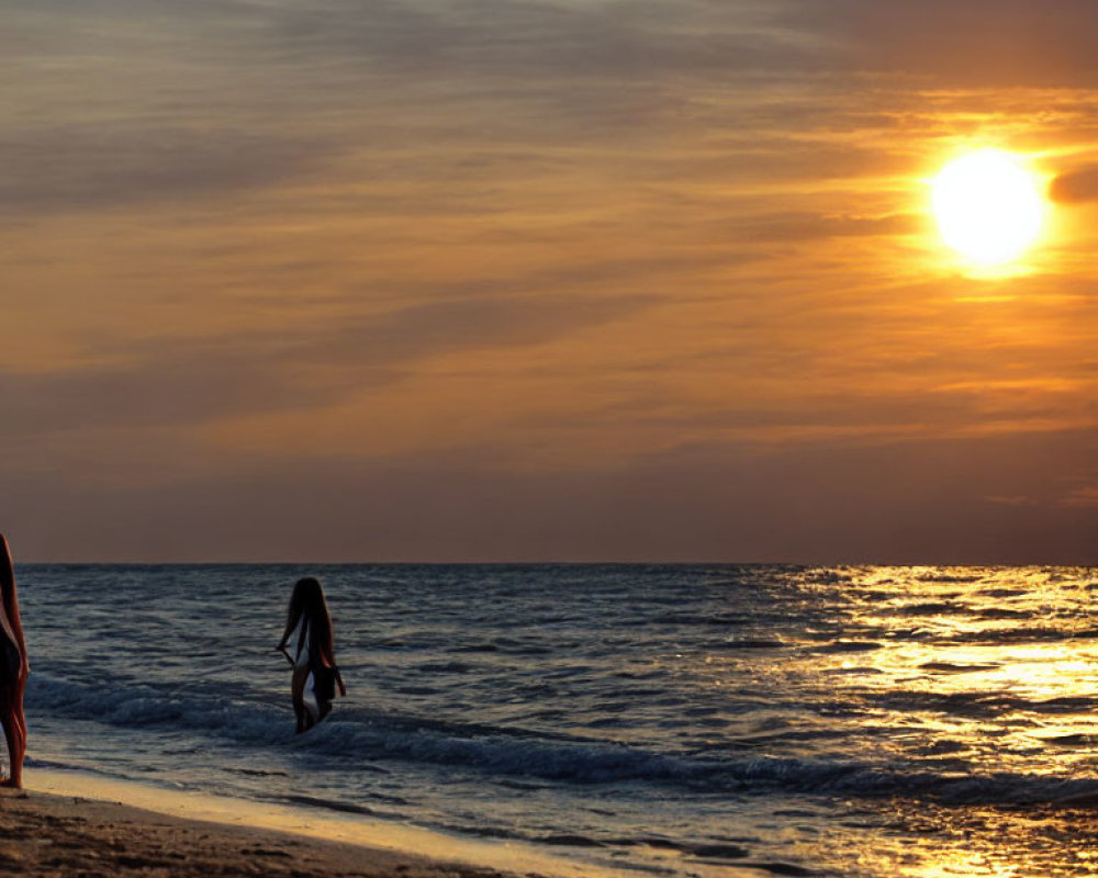 Person walking along shore at sunset with sun reflecting on ocean waves.