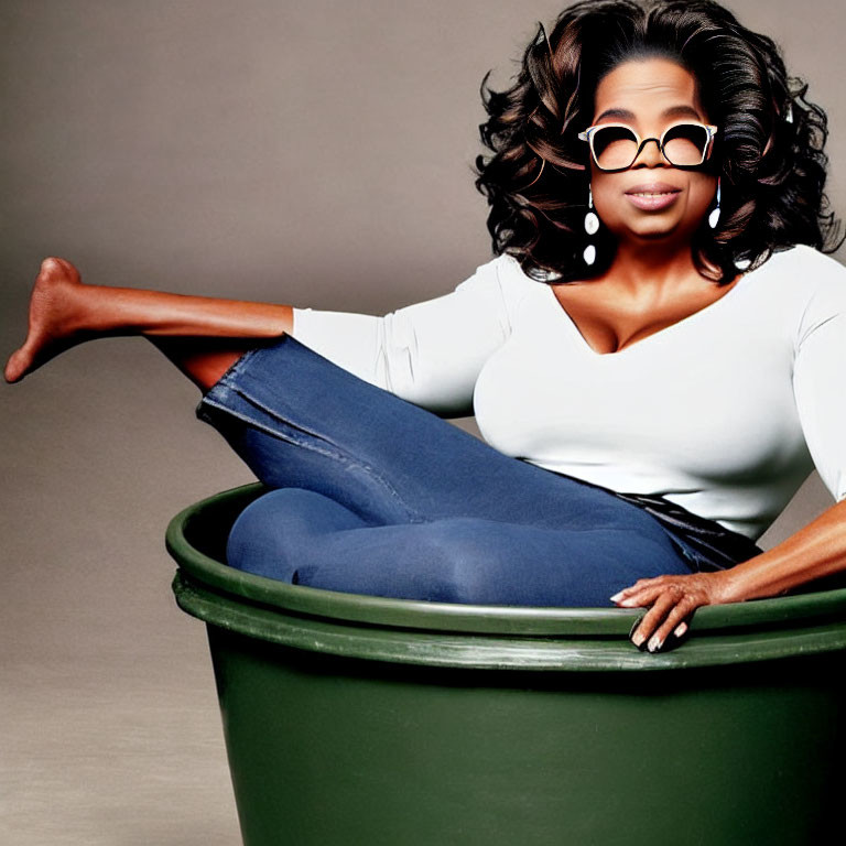 Person in sunglasses sits inside green trash can with white top and blue jeans