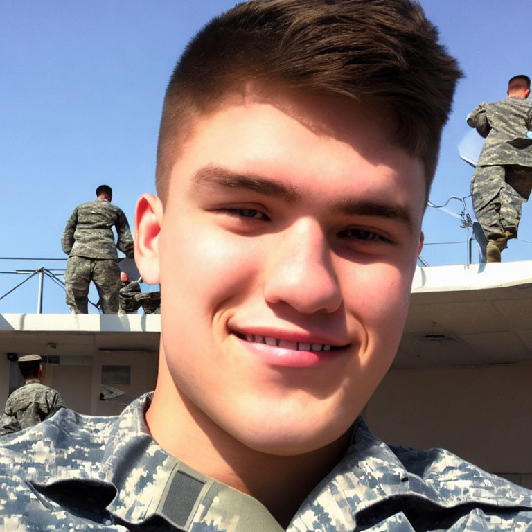 Young man in military uniform smiling with others in background under clear skies