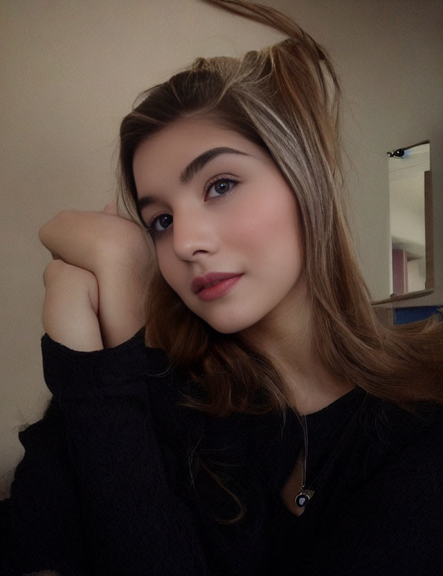 Young woman with fair skin and dark hair in black top and pendant necklace poses with hand on chin