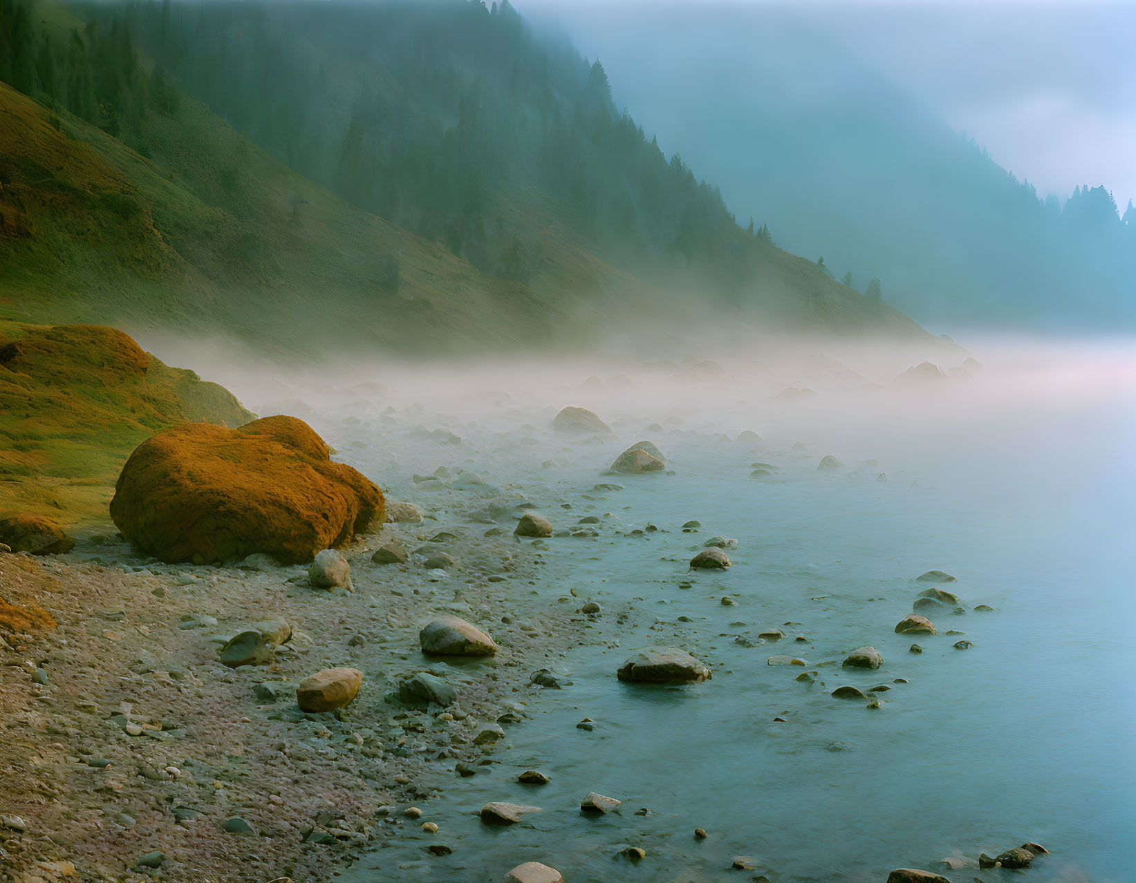 Tranquil lakeside scene with moss-covered boulders and foggy mountain backdrop