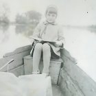 Vintage-dressed woman reading book in rowboat with ship backdrop