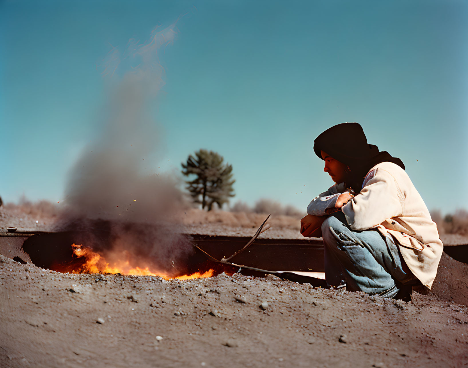 Person squatting by open flame in hoodie and jeans under clear blue sky