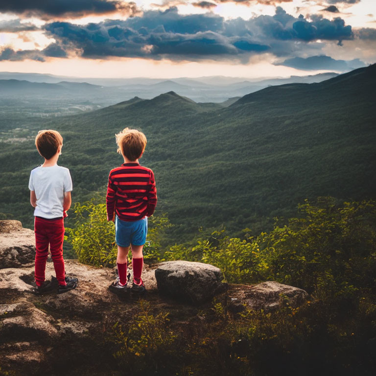 Children on rocky outcrop overlooking valley with mountains under dramatic sunset sky