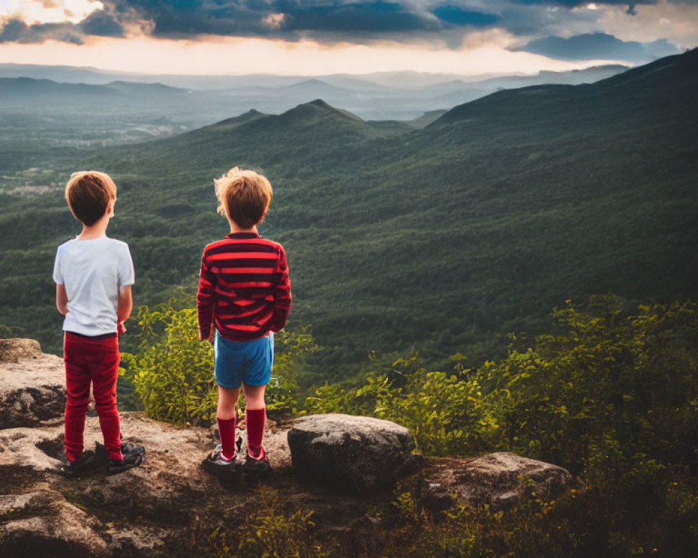 Children on rocky outcrop overlooking valley with mountains under dramatic sunset sky